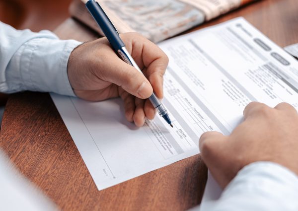 Person reviewing and writing on financial documents with a pen on a wooden desk.